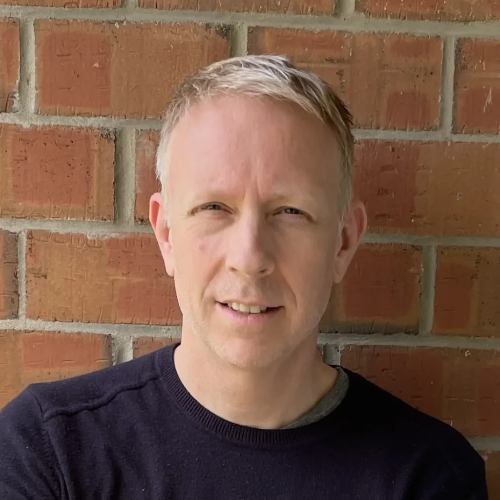 Headshot of author G.S. Arnold wearing a dark purple t-shirt, standing against a brick wall